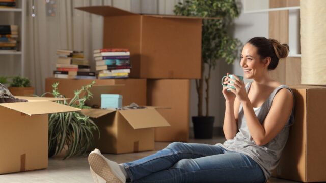 Relaxed woman holding a cup of tea, sitting on the floor surrounded by moving boxes
